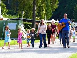 Mann mit Gitarre und einige Kinder gehen gemeinsam über einen Zeltplatz
